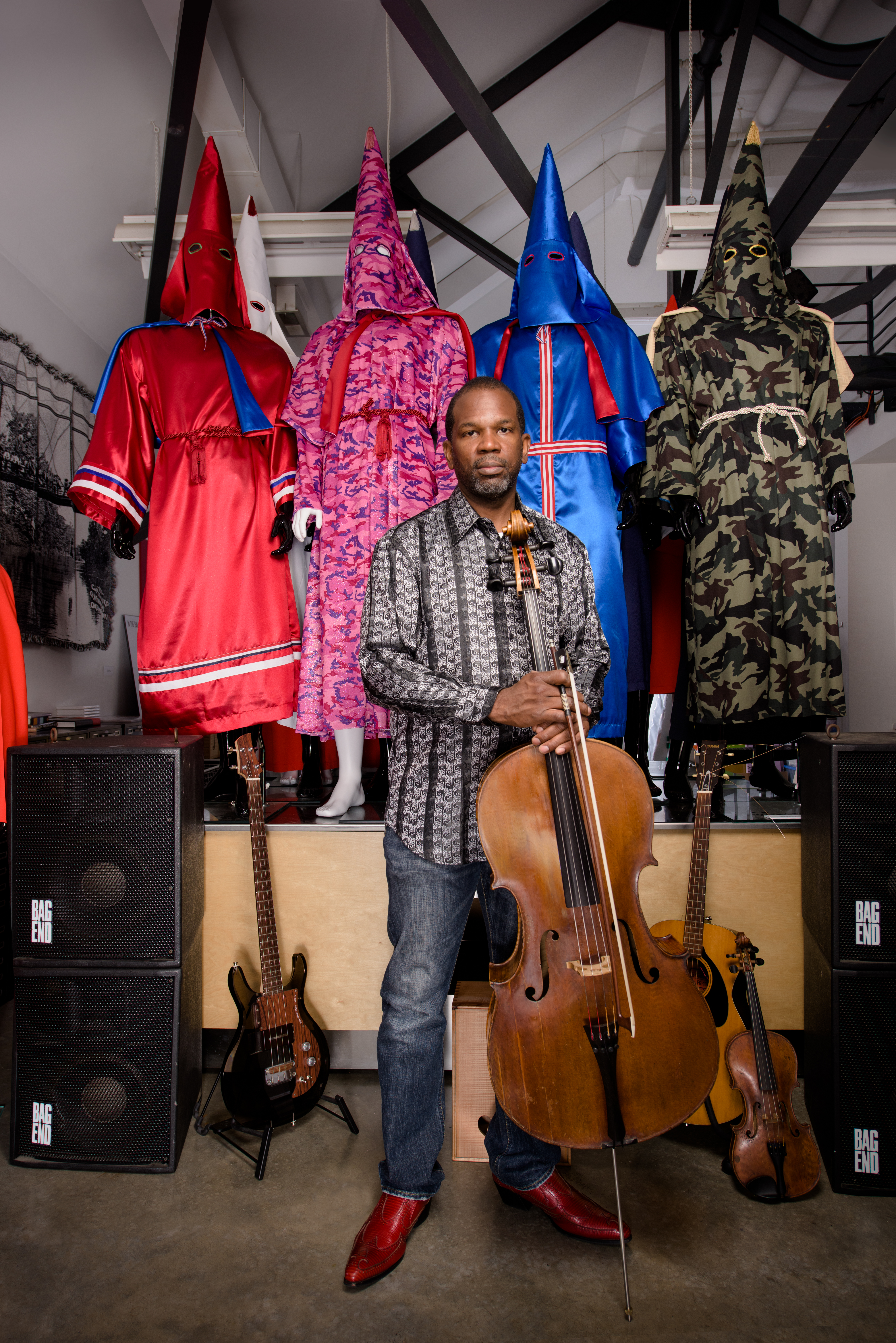 Paul Rucker standing with his cello in front of his art pieces, KKK robes reimagined with multicolored cloth
